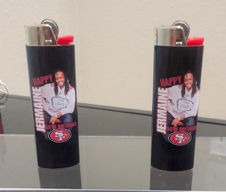 May include: Two black lighters with red tops. Each lighter has a personalized label with a photo of a man, the text "Jermaine", "Happy 50th Birthday", and a red and white logo.