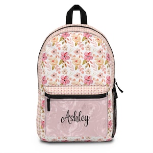 May include: A floral backpack with a pink and white pattern featuring roses and other flowers. The backpack has a black zipper and a front pocket with the name "Ashley" in cursive. The bag has a black handle and black mesh side pocket.