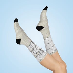 May include: A pair of novelty socks with a light beige background and black toes and heels. The socks feature a white design resembling belts and buckles. The socks are displayed against a light blue background.