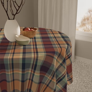 Puede incluir: Un mantel redondo con un estampado de cuadros en tonos azules, marrones y naranjas. Un jarrón blanco con ramas decorativas, una pequeña cesta de manzanas y un cuenco están sobre la mesa. El mantel cubre toda la mesa.