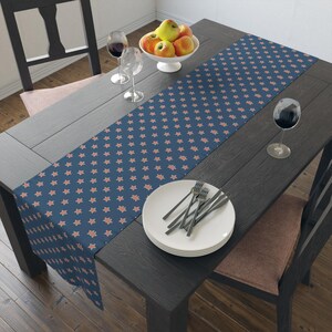 May include: A navy blue table runner featuring a repeating pattern of coral stars. The runner is laid on a dark wooden table, set with plates, cutlery, and wine glasses. A bowl of apples is in the center.
