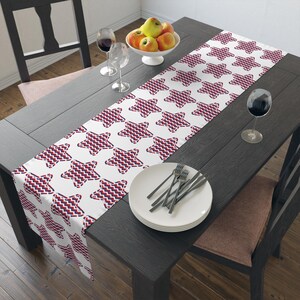 May include: A white table runner featuring a repeating pattern of red and navy blue stars. The runner is laid on a dark wooden table, with a white bowl of apples, wine glasses, and a plate with cutlery.