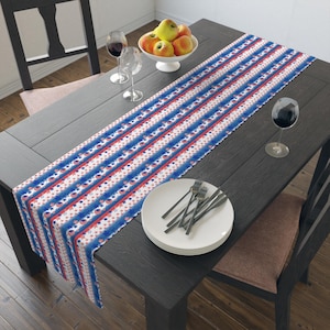 May include: A table runner with a repeating pattern of red and blue stripes and stars. The runner is placed on a dark wooden table, with a white bowl of apples, wine glasses, and plates with forks.
