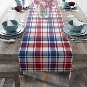 May include: A dining table set with a red, white, and blue tartan table runner. Teal bowls, white plates, wine glasses, and cutlery are arranged. A vase of pink flowers is in the center of the table.