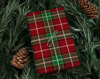 May include: A wrapped gift box with a red, green, and white plaid pattern, tied with twine. The box is surrounded by green pine needles and pine cones, creating a festive holiday theme.