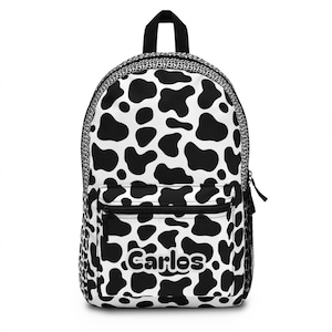 May include: A black and white backpack with a cow print design. The backpack has a front pocket, a top handle, and the name "Carlos" printed on the front.