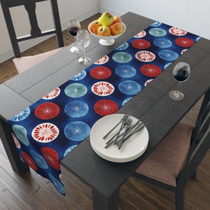 May include: A navy blue table runner featuring a pattern of red, white, and blue circular designs. The runner is on a dark grey wooden table. A white bowl of fruit and wine glasses are also visible on the table.