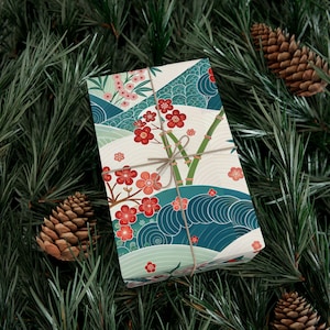 May include: A wrapped gift with a Japanese-inspired design featuring red cherry blossoms, bamboo stalks, and blue wave patterns. The gift is tied with twine and rests on a bed of green pine needles, with pine cones scattered around.