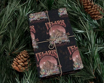 May include: A wrapped gift with a dark background featuring the word "VELARIS" in a stylized font. The design includes a mountain scene with floral and butterfly accents. The gift is tied with twine and rests on a bed of pine needles and pine cones.