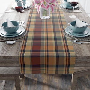 May include: A plaid table runner in shades of brown, orange, and green, displayed on a wooden table. The runner is the focal point, with a repeating pattern of intersecting lines. The table is set with plates, bowls, silverware, and wine glasses.