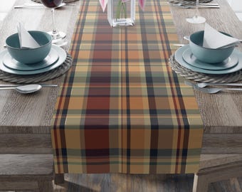 May include: A plaid table runner in shades of brown, orange, and green, displayed on a wooden table. The runner is the focal point, with a repeating pattern of intersecting lines. The table is set with plates, bowls, silverware, and wine glasses.