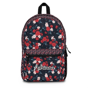 May include: A floral backpack with a navy blue background and a pattern of red and white roses. The backpack has a black zipper and a black handle. The name "Adriana" is written in cursive on the front.