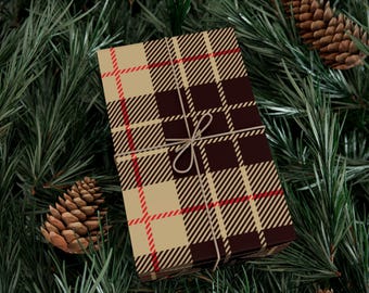 Puede incluir: Una caja de regalo envuelta con un patrón de cuadros marrón y beige, acentuada con líneas rojas, está atada con hilo. El regalo descansa sobre un lecho de agujas de pino verdes y está acompañado de piñas, creando un tema festivo de vacaciones.
