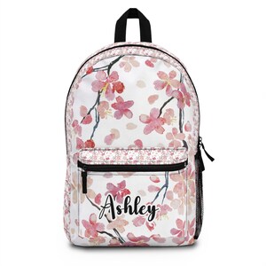 May include: White backpack with a floral cherry blossom design in pink and red. The backpack has a black zipper and a front pocket. The name "Ashley" is written in cursive on the front.