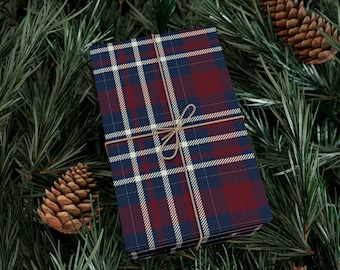 May include: A gift wrapped in plaid paper with a navy blue, burgundy, and white pattern, tied with twine. The gift rests on a bed of green pine needles, with pine cones scattered around.
