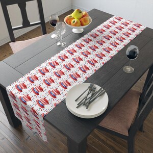 May include: A white table runner featuring a repeating pattern of red, white, and blue Uncle Sam hats and confetti. The runner is laid on a dark wooden table, set with plates, cutlery, and wine glasses.