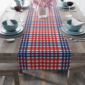 Pode incluir: Um caminho de mesa xadrez vermelho, branco e azul marinho é exibido em uma mesa de madeira, pronto para uma refeição. O caminho de mesa apresenta um padrão xadrez clássico. A mesa está posta com pratos, tigelas, talheres e taças de vinho.