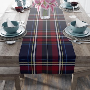 May include: A navy blue tartan table runner with red, white, and yellow detailing. The runner is laid on a wooden table, set for a meal with plates, cutlery, and wine glasses. A vase of pink flowers is in the centre.