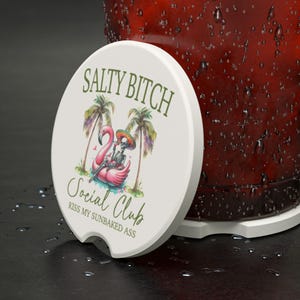 May include: White round car coaster with the text "Salty Bitch Social Club" and a flamingo skeleton design. The flamingo is wearing a sombrero and is floating on a pink inflatable. Palm trees are on either side of the flamingo.