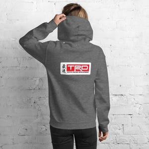 May include: A gray hooded sweatshirt with the TRD logo on the back. The logo is a white rectangle with the letters "TRD" in red and Japanese characters above. The words "TOYOTA RACING DEVELOPMENT" are below the logo.