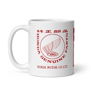 May include: White ceramic mug featuring a red Honda logo with the text "HONDA GENUINE PARTS" and Japanese characters. The mug has a curved handle and a cylindrical shape, perfect for coffee or tea.