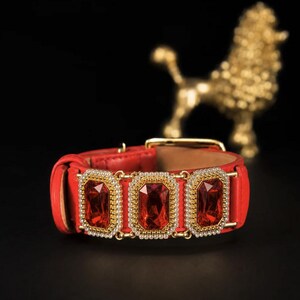 Könnte beinhalten: Ein rotes Lederhalsband mit drei rechteckigen, facettierten roten Edelsteinen in goldfarbenen Metallfassungen. Jeder Edelstein ist von einem Rand aus kleinen, klaren weißen Kristallen umgeben. Das Halsband hat eine goldfarbene Metallschnalle.