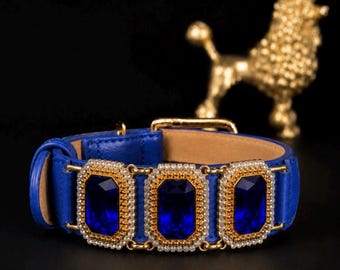 Puede incluir: Un collar de cuero azul real con tres grandes piedras preciosas rectangulares de color azul oscuro facetado. Cada piedra está enmarcada por un borde de pequeños cristales brillantes engastados en metal dorado. El collar incluye una hebilla dorada.