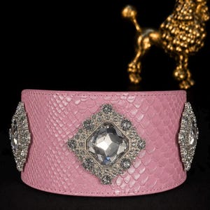 May include: A wide pink cuff bracelet featuring a textured snakeskin pattern. The center is adorned with a large, square-cut clear rhinestone surrounded by smaller pavé crystals in a diamond-shaped setting, flanked by two matching crystal accents.