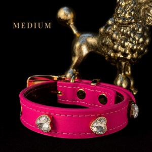 Puede incluir: Un collar rosa brillante con cristales transparentes en forma de coraz&oacute;n engastados en metal dorado a lo largo de la correa. El collar incluye una hebilla y herrajes dorados, con la palabra MEDIUM impresa en blanco sobre un fondo negro.