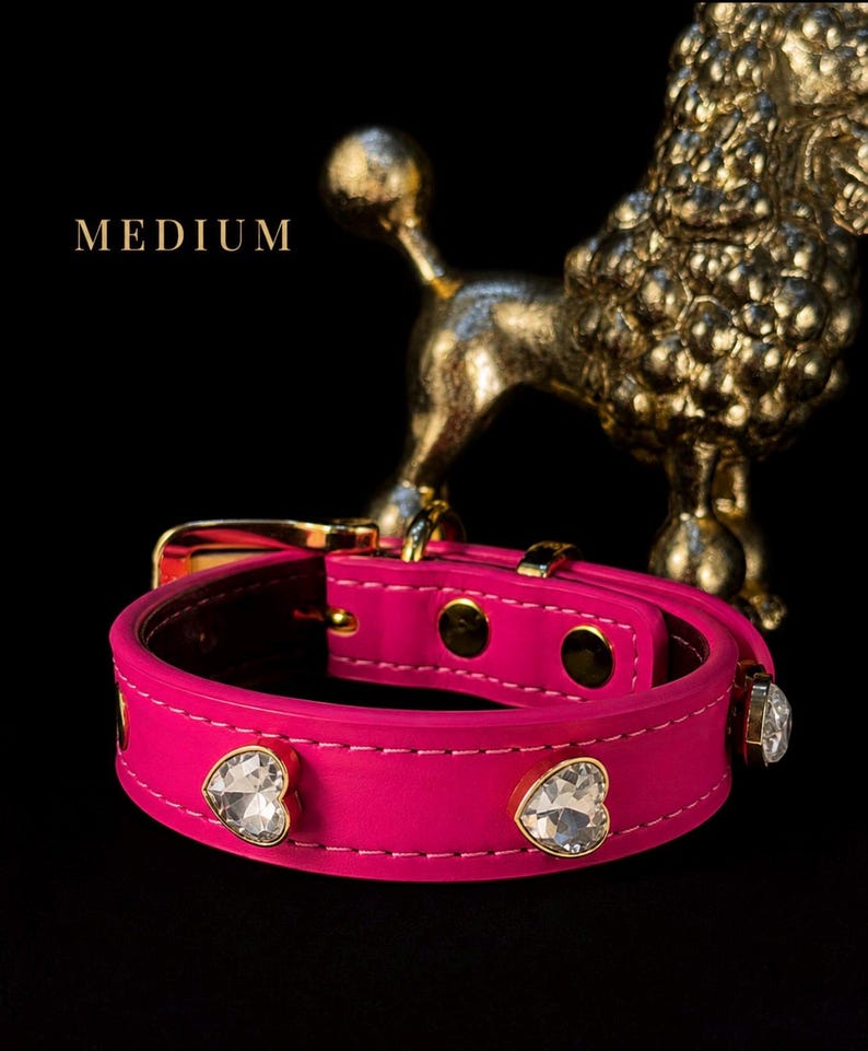 Puede incluir: Un collar rosa brillante con cristales transparentes en forma de coraz&oacute;n engastados en metal dorado a lo largo de la correa. El collar incluye una hebilla y herrajes dorados, con la palabra MEDIUM impresa en blanco sobre un fondo negro.
