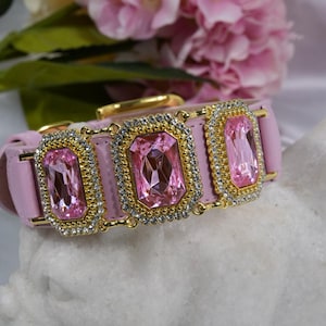 Op de afbeelding: Roze hondenhalsband met gouden accenten en drie grote, rechthoekige roze edelstenen. De edelstenen zijn omgeven door een rand van kleine, heldere kristallen. De achtergrond toont roze bloemen.