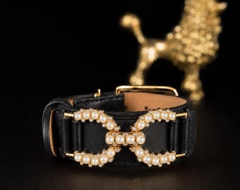Puede incluir: Una pulsera de cuero negro con un adorno central de hebilla de metal dorado en forma de doble bucle. El adorno está decorado con pequeñas perlas blancas artificiales y cristales transparentes. La pulsera incluye un cierre de hebilla de metal dorado.