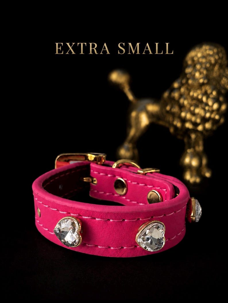Puede incluir: Un collar para mascota de color rosa intenso con cristales transparentes en forma de coraz&oacute;n, herrajes de metal dorado y costuras blancas. El texto EXTRA SMALL aparece sobre el collar, que se encuentra sobre un fondo negro.
