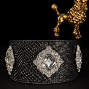 Könnte beinhalten: Ein breites schwarzes Armband mit einer strukturierten Schlangenhautoptik. Das Armband ist mit drei rautenförmigen silberfarbenen Fassungen verziert, die jeweils mit klaren Kristallen um einen großen, facettierten Mittelstein besetzt sind.