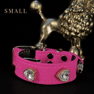 Puede incluir: Un collar de color rosa brillante con una hebilla plateada y dos adornos de cristal transparente en forma de coraz&oacute;n sujetos con monturas met&aacute;licas. La palabra SMALL est&aacute; impresa en blanco sobre el collar, situado frente a una figura dorada.