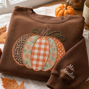 May include: A brown sweatshirt featuring a large, multi-patterned pumpkin appliqué on the front with sections of leopard print, floral, orange gingham, and polka dots. The sleeve is embroidered with the name Ms. Amelia in a cursive script.