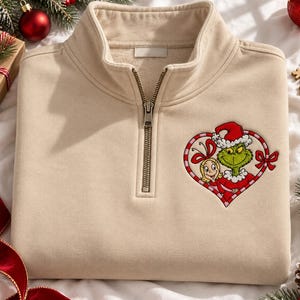 Può includere: Una felpa beige con zip a un quarto e un ricamo sul petto sinistro. Il disegno raffigura un personaggio verde e una bambina bionda all'interno di una cornice a forma di cuore a righe rosse e bianche, entrambi con cappelli natalizi.