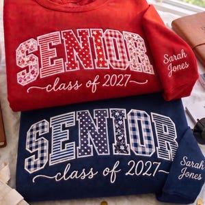 May include: Two crewneck sweatshirts, one red and one navy blue, each featuring the word SENIOR in large block letters with varied patterns and the text class of 2027 embroidered below. The sleeve of each sweatshirt is personalised with Sarah Jones.