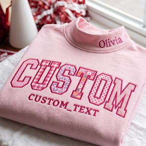 May include: A light pink mock neck sweatshirt featuring the embroidered name Olivia on the collar. The front displays the word CUSTOM in large varsity-style letters with various pink patterned fills, and the text CUSTOM_TEXT below it.