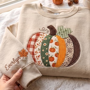 Peut inclure: Un sweat-shirt beige à col rond orné d'une grande applique en forme de citrouille composée de divers tissus à motifs orange, verts et marron. La manche est brodée du prénom Everlyn accompagné d'un motif de feuille.