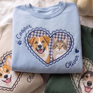 May include: Three crewneck sweatshirts in light blue, beige, and dark green, each featuring an embroidered heart-shaped frame with a blue and white gingham pattern. The designs include detailed portraits of dogs and a cat, with names like Cooper and Oliver stitched in blue thread alongside small paw print and heart icons.
