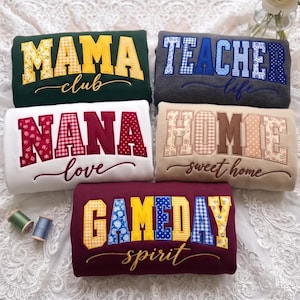 May include: Five folded sweatshirts featuring fabric applique letters and cursive text. The shirts display the phrases MAMA club in green, TEACHER life in gray, NANA love in white, HOMIE sweet home in beige, and GAMEDAY spirit in maroon.