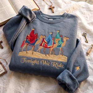 Puede incluir: Una sudadera de cuello redondo color azul pizarra con un bordado de tres figuras con coronas montadas en camellos sobre un paisaje desértico. El texto "Tonight We Ride" está bordado bajo los camellos y una pequeña estrella dorada en la manga.