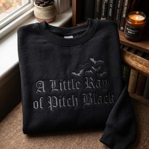 May include: Black crewneck sweatshirt with the phrase "A Little Ray of Pitch Black" embroidered in gray gothic lettering. Above the text are embroidered bats. The sweatshirt is displayed on a brown chair.