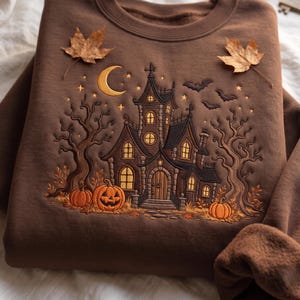 May include: A brown crewneck sweatshirt featuring an embroidered design of a haunted house, bare trees, pumpkins, a crescent moon, and bats. The scene includes glowing yellow windows and stars, with two dried maple leaves placed on the fabric.