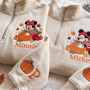 May include: Two beige quarter-zip sweatshirts featuring embroidered designs. One displays Minnie Mouse with a pumpkin and the name Minnie, the other Mickey Mouse with a pumpkin and the name Mickey. Both sleeves feature an embroidered pumpkin and letter P.
