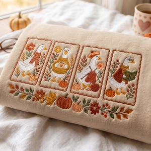 Op de afbeelding: Een beige sweatshirt met vier rechthoekige geborduurde panelen, elk met een witte gans in herfstkleding. De ganzen dragen sjaals, truien en een bril, omringd door pompoenen, bloemen en bladeren in warme oranje en bruine tinten.