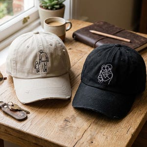 May include: Two baseball caps with embroidered designs sit on a wooden table. One is tan with a father and child design, the other is black with an embracing couple. Other items include sunglasses, keys, a notebook, and a coffee mug.