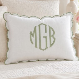 Op de afbeelding: Een rechthoekig wit sierkussen met een geschulpte rand afgewerkt in saliegroen. Het midden van het kussen toont een groot, geborduurd monogram met de letters MGB in saliegroene draad, passend bij het detail van de geschulpte rand.