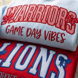 Op de afbeelding: Een lichtblauw sweatshirt met de rood-wit gestreepte tekst WARRIORS met rode stippen-strikken, en de rode tekst GAME DAY VIBES. Daaronder een rood sweatshirt met de blauw-wit gestreepte tekst LIONS met blauwe stippen-strikken.